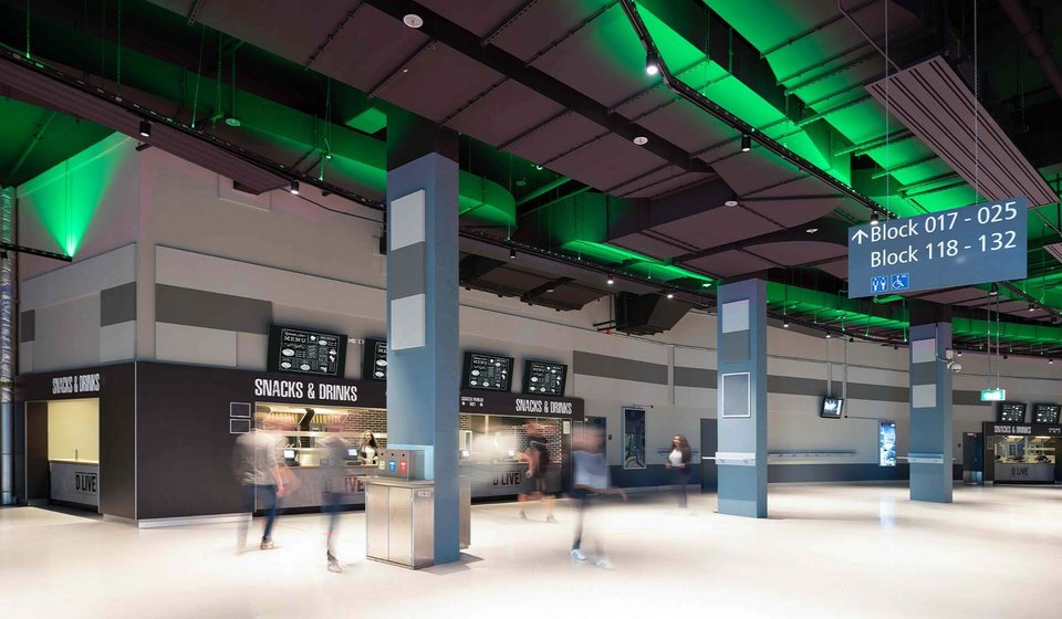 Concourse in PSD BANK DOME. The view falls on a stand with the signage “Snacks & Drinks”. Above the signage there are several display boards with the food and drink menu. The ceiling glows green. 